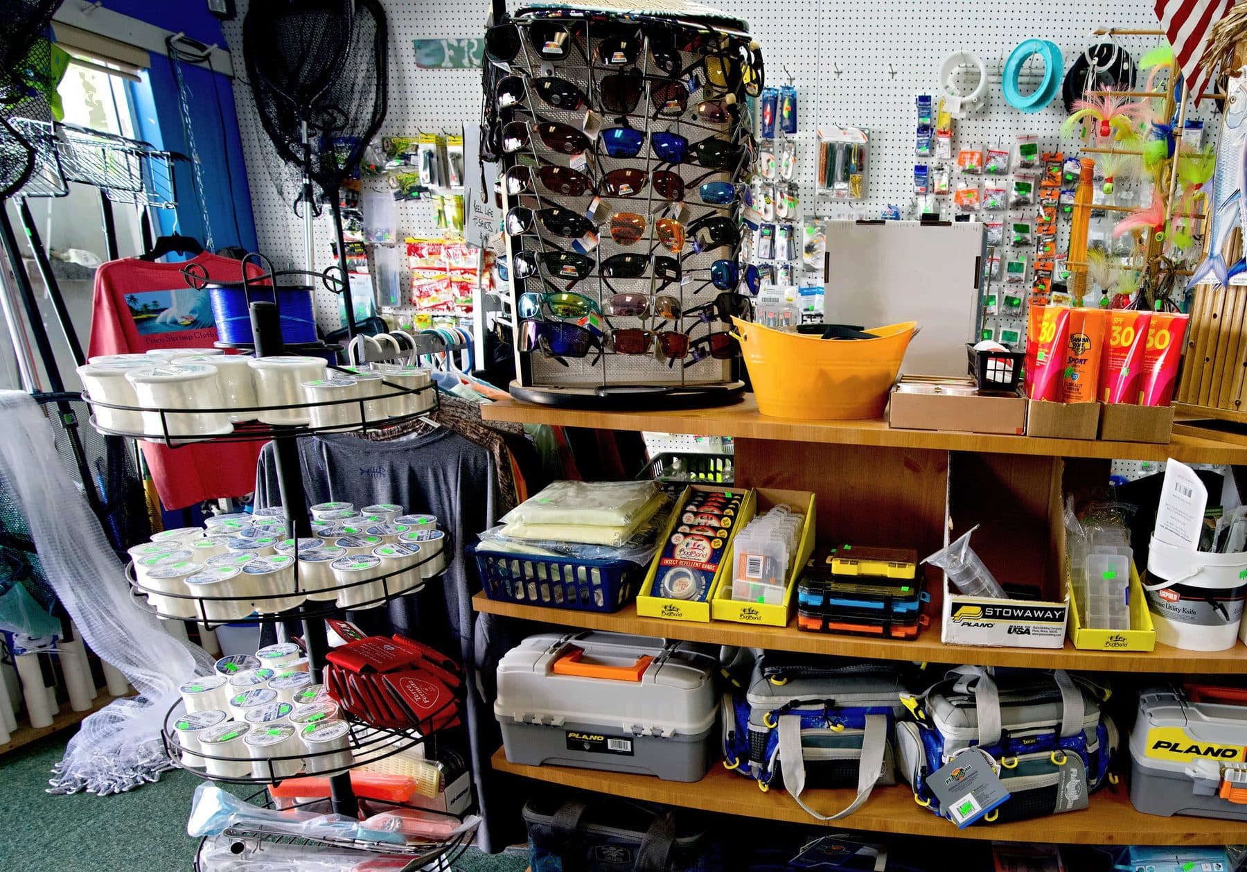 Curved wooden shelf of Plano tackle boxes and Flambeau tackle bags under a sunglasses rack, a tiered line spool rack at left